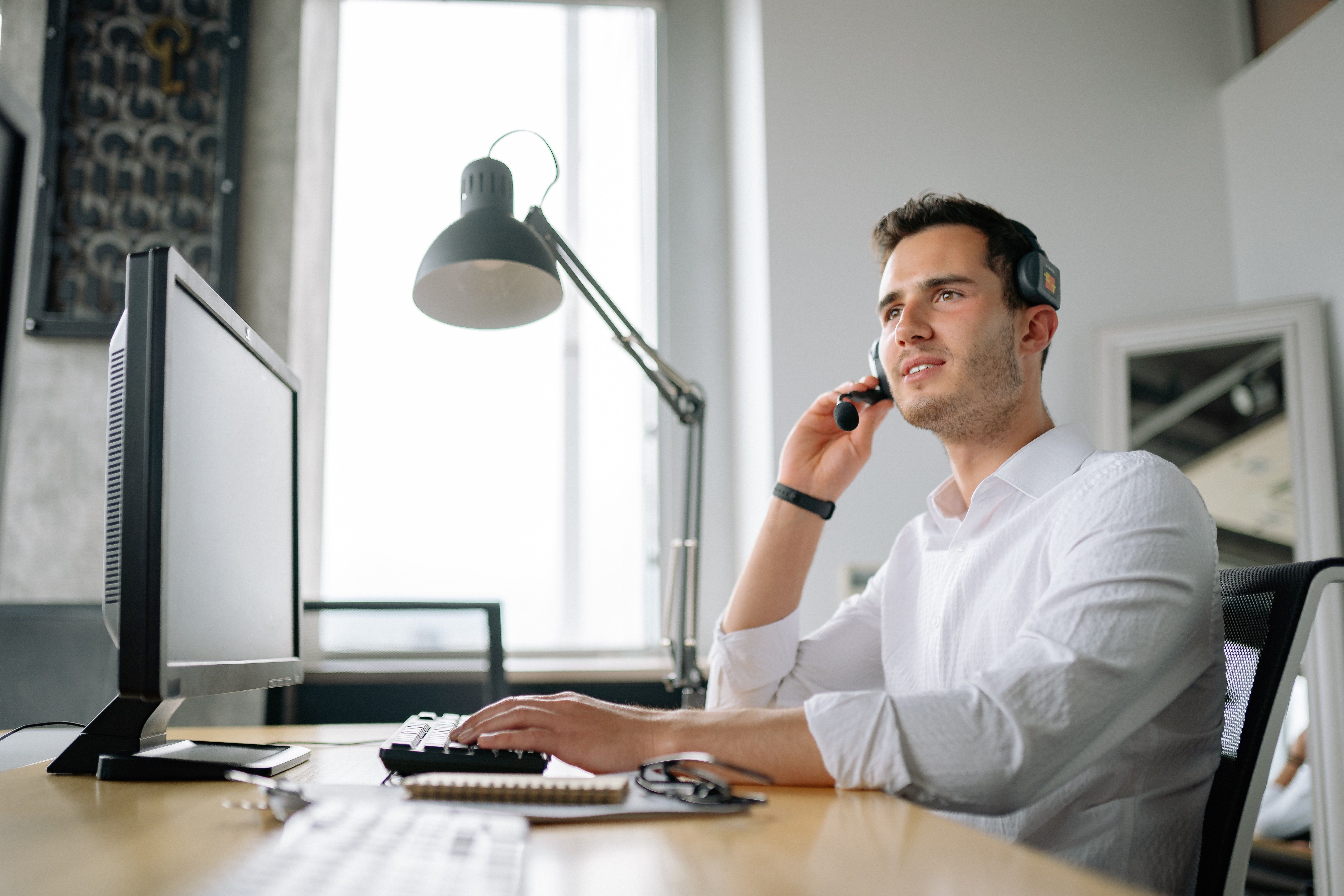 A man in a white shirt sitting at a desk wearing a headset and speaking to someone while he types on a computer keyboard.